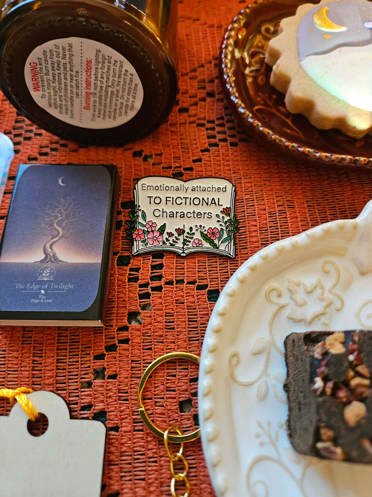 A styled flat-lay featuring a silver enamel pin with a floral design and the text 'Emotionally attached TO FICTIONAL Characters' on a textured orange table runner, surrounded by cozy props including a candle, a cookie, and a sugar cube.