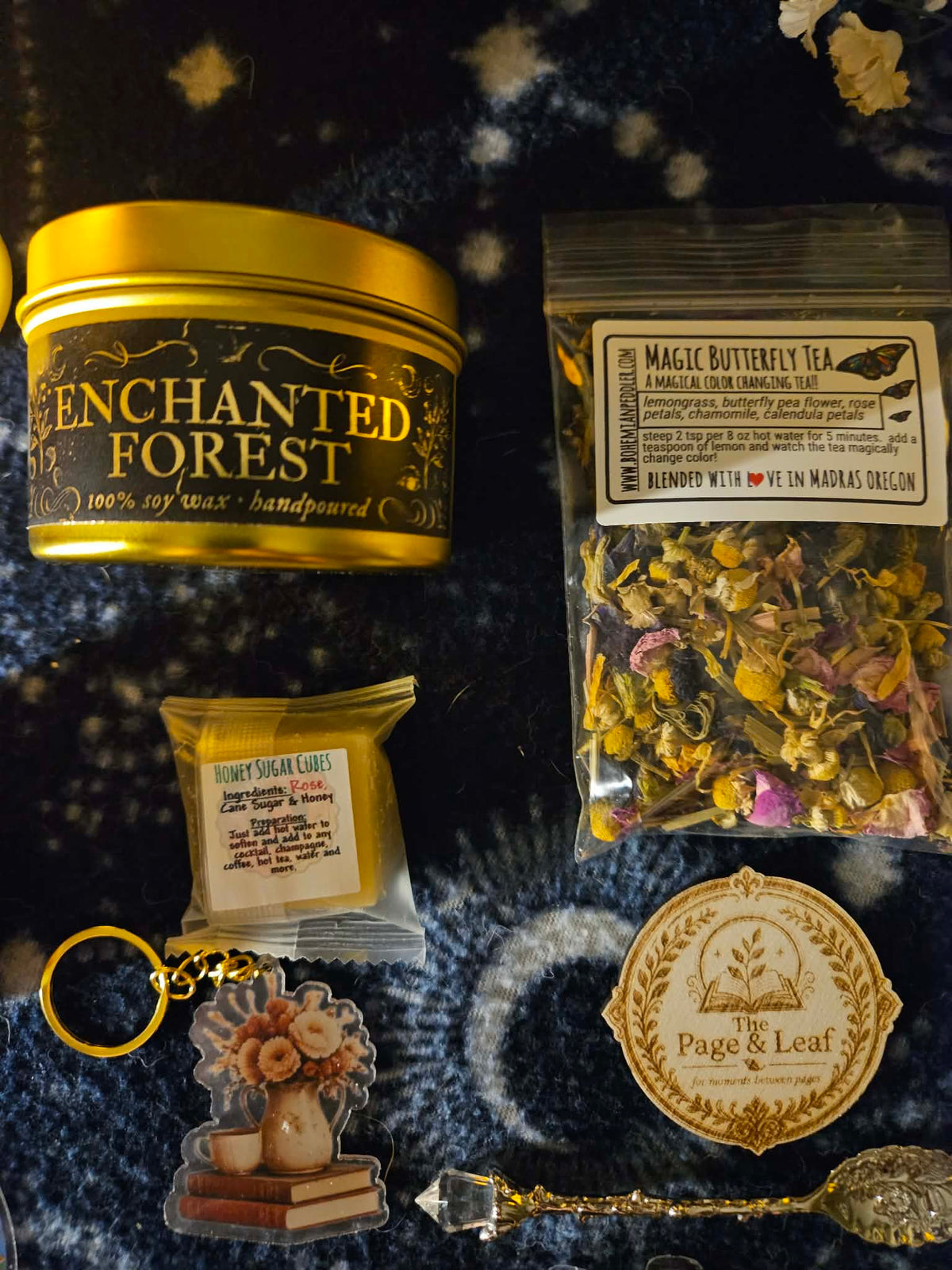 A flat-lay collection of handmade products on a dark blue starry blanket, including an Enchanted Forest wax melt tin, a bag of Magic Butterfly Tea, a honey square keychain, a page & leaf coaster, and a crystal spoon.