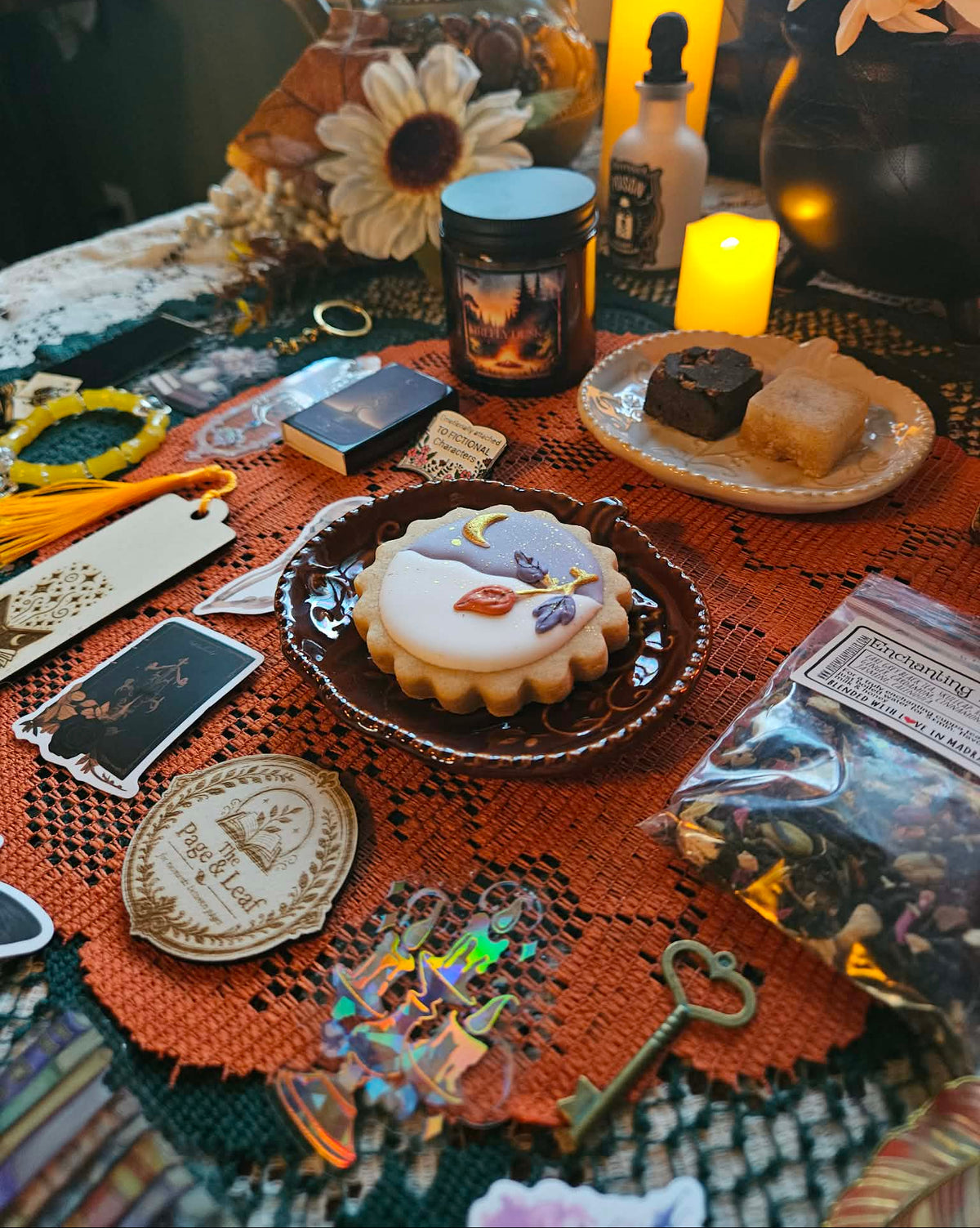 A styled tabletop display with a decorated sugar cookie in a dark glass dish, surrounded by candles, tarot cards, a heart-shaped key, crystals, and other mystical accessories on an orange lace runner.