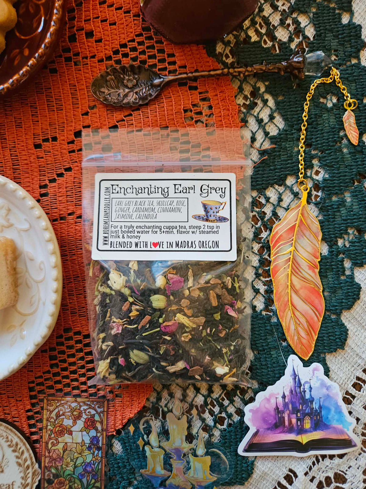 A clear plastic bag of Enchanted Earl Grey tea blend sits on a patterned orange and green textile with decorative props, including a spoon, a feather bookmark, and a castle sticker.