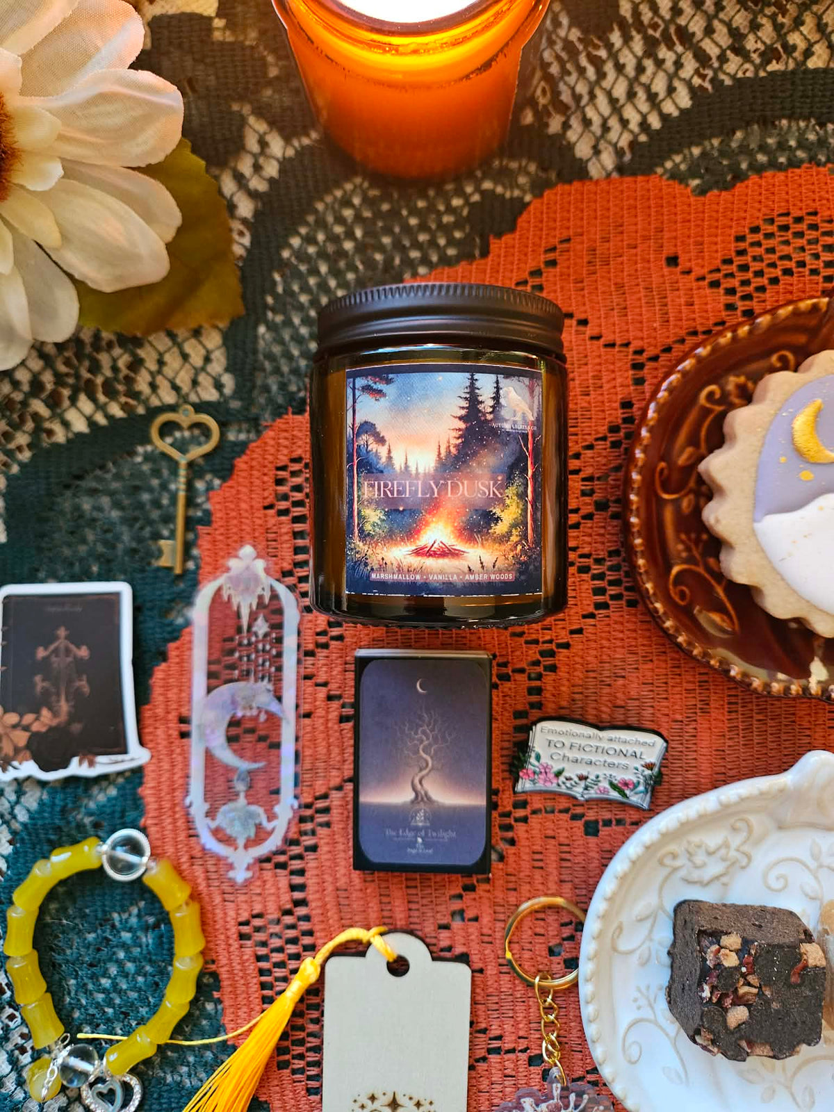 A styled flat-lay featuring a 'Firefly Dusk' scented candle in a dark glass jar with a black lid, surrounded by mystical and cozy props including stickers, a white crescent moon bookmark, and  a yellow beaded bracelet all arranged on an orange patterned textile.