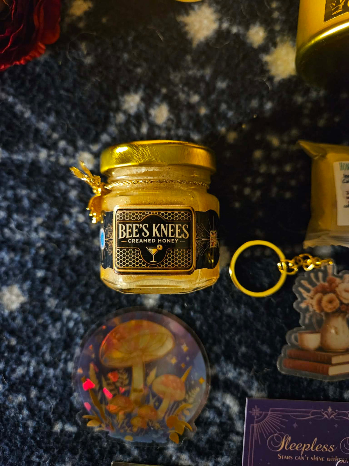 A small glass jar of BEE'S KNEES Creamed Honey with a gold lid and label, displayed on a dark textured surface alongside other themed items including a mushroom coaster, keychain, and card.