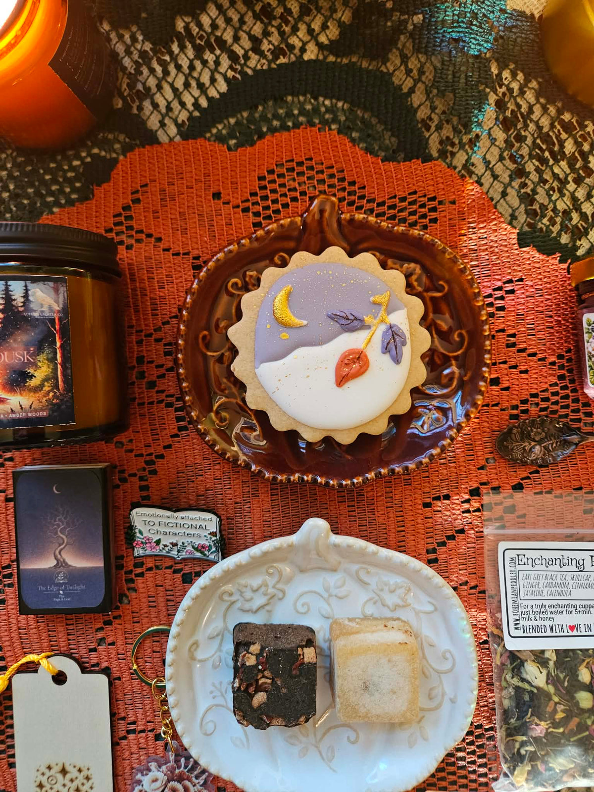 A styled flat-lay of autumn-themed products on a patterned textile background, featuring a decorated sugar cookie on an ornate brown plate, a white plate with two sugar cubes, a candle jar, a match box, and an enamel pin.