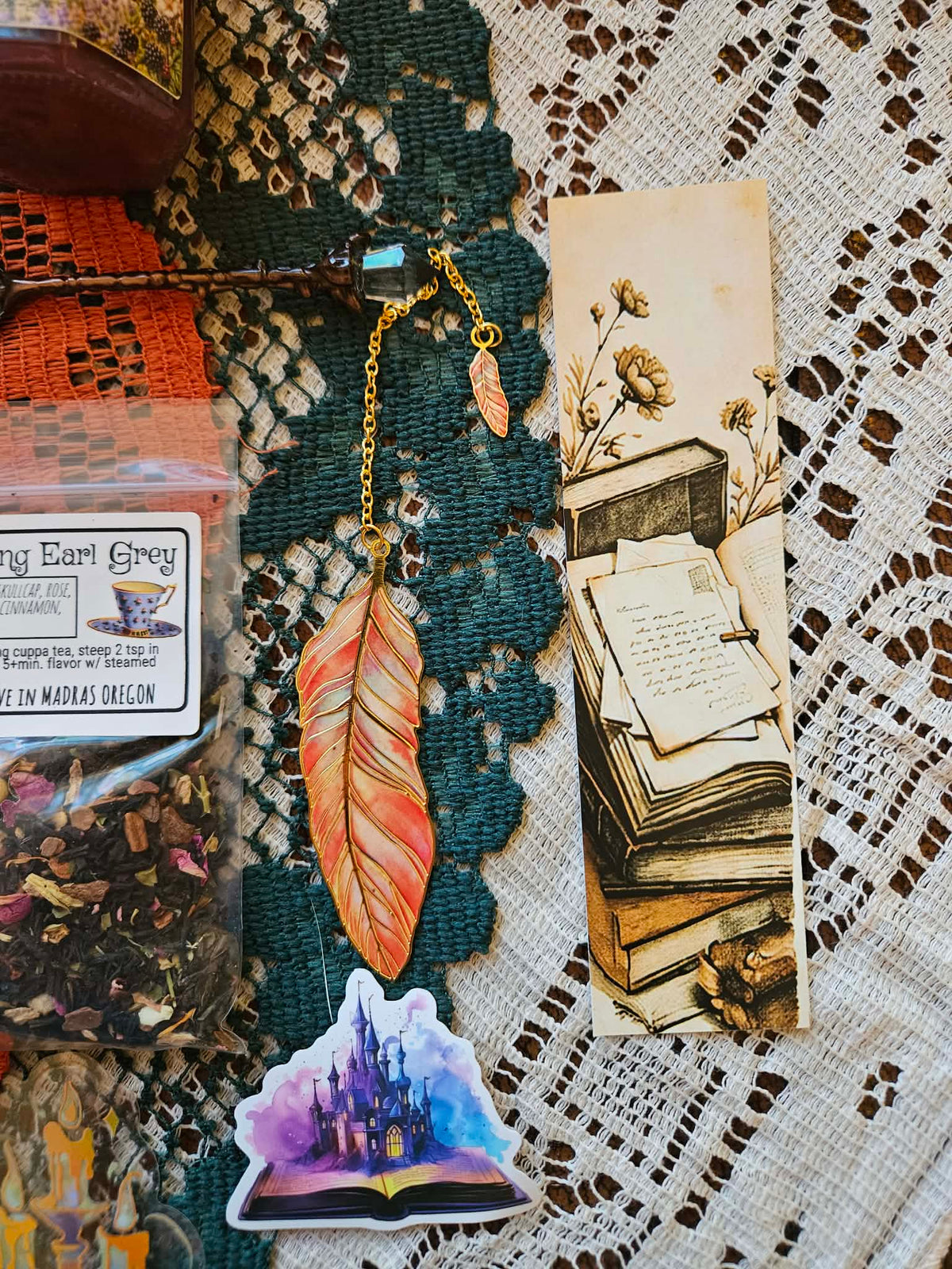 A styled flat-lay of bohemian-themed stationery and accessories, featuring a paper bookmark with an illustration of old books, a colorful feather bookmark, a purple castle sticker, dried flowers, and a tea bag on a lace doily.