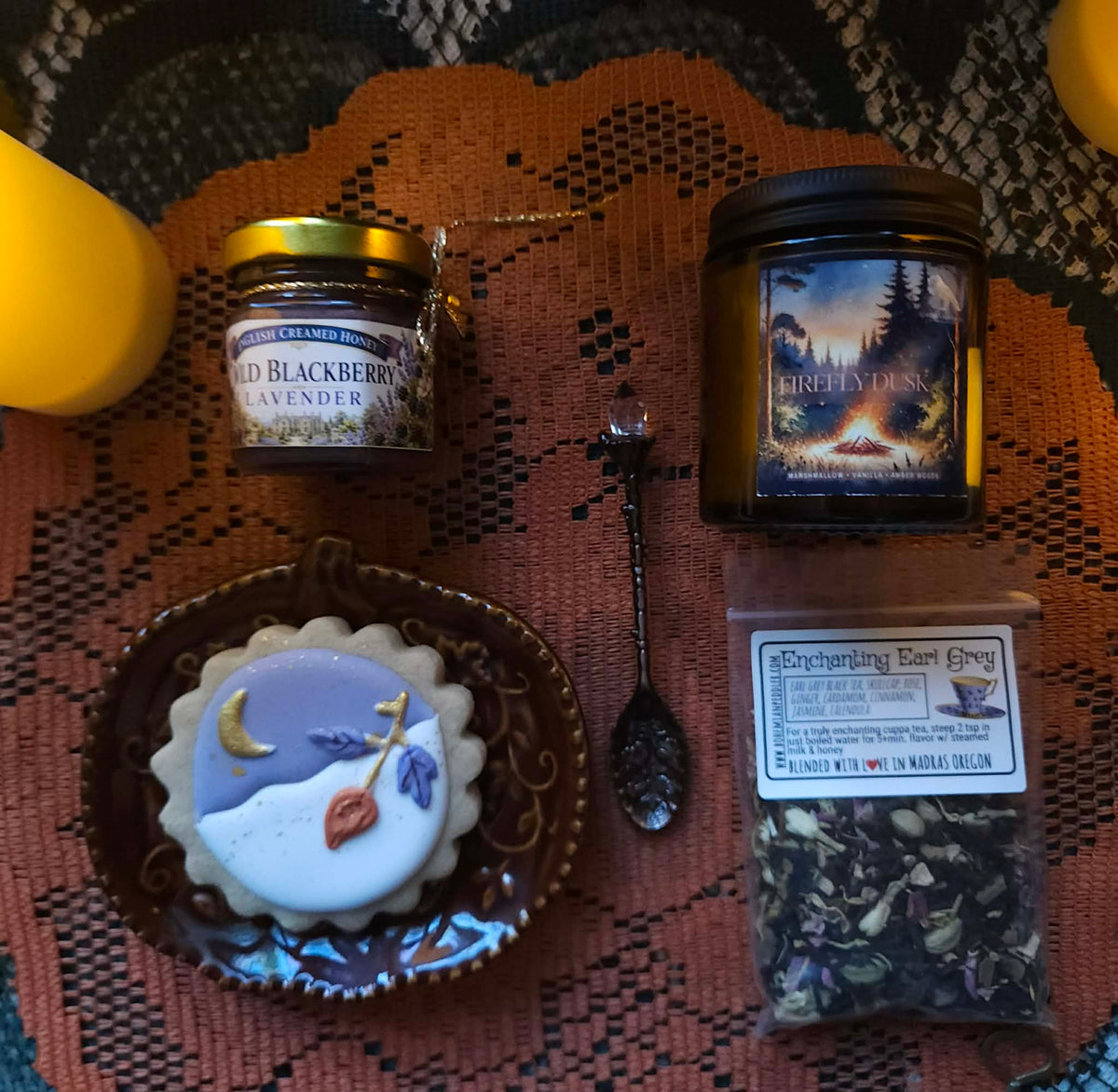 A cozy flat-lay arrangement of tea and a cookie on a patterned rust-orange textile. Visible products include a jar labeled Wild Blackberry Lavender Tea, a candle labeled Firefly Dusk, a small decorative plate with a moon-and-stars cookie, and a bag of Enchanted Earl Grey Tea.