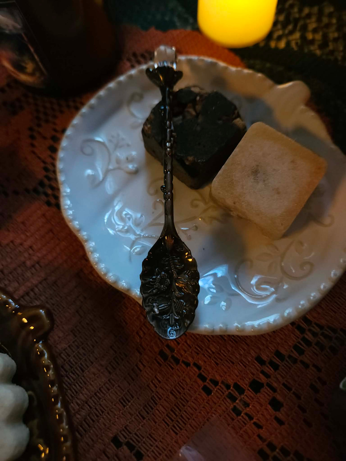 A decorative white ceramic plate with embossed scrollwork, holding a dark spoon and two sugar cubes, set on a brown patterned tablecloth with a warm yellow light in the background.