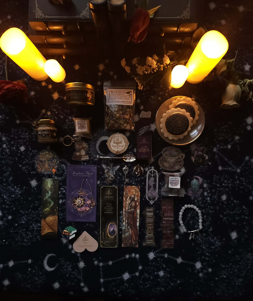 A styled flat-lay of various witchcraft and occult accessories arranged on a dark starry fabric background, illuminated by two glowing yellow pillar candles. The collection includes tarot cards, crystals, a brass incense burner, a silver athame, a wooden wand, a heart-shaped card, and other mystical items.