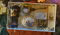 A curated gift box set containing a candle, a jar of blackberry lavender honey, a matchbox, a decorative cookie, a bag of loose herbal tea, a demitasse spoon, and a wooden bookmark, all arranged in cream tissue paper inside a white box.