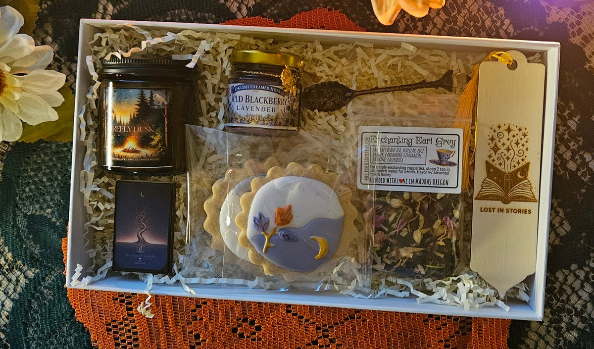 A curated gift box set containing a candle, a jar of blackberry lavender honey, a matchbox, a decorative cookie, a bag of loose herbal tea, a demitasse spoon, and a wooden bookmark, all arranged in cream tissue paper inside a white box.