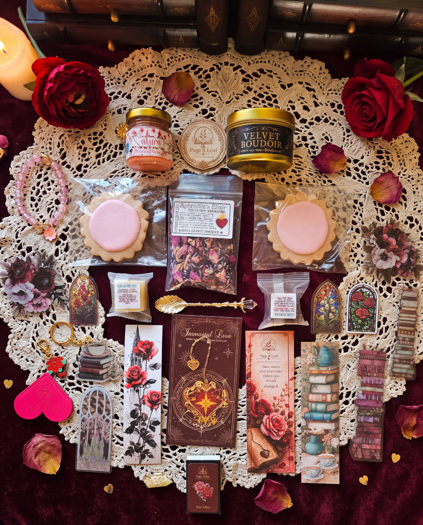A styled flat-lay of a curated gift basket of tea and snack assortment arranged on a cream lace doily over a dark burgundy fabric. Visible items include two pink macarons, a jar labeled Velvet Bontrio, a small jar labeled Rose Petal Tea, assorted packaged snacks, decorative cards, bookmarks, a gold spoon, rose petals, and red roses.