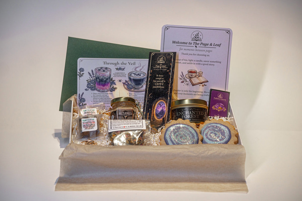 A curated gift basket containing multiple packaged items arranged in a clear plastic tray lined with cream tissue paper. Visible contents include printed cards, a dark box, a small jar, round packaged items, and two round blue-and-white wrapped items, all set against a plain light background.