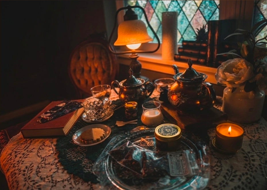 A cozy, dimly lit tabletop scene with a teapot, candle, book, and other warm-toned objects arranged on a lace-covered table near a window and lamp.